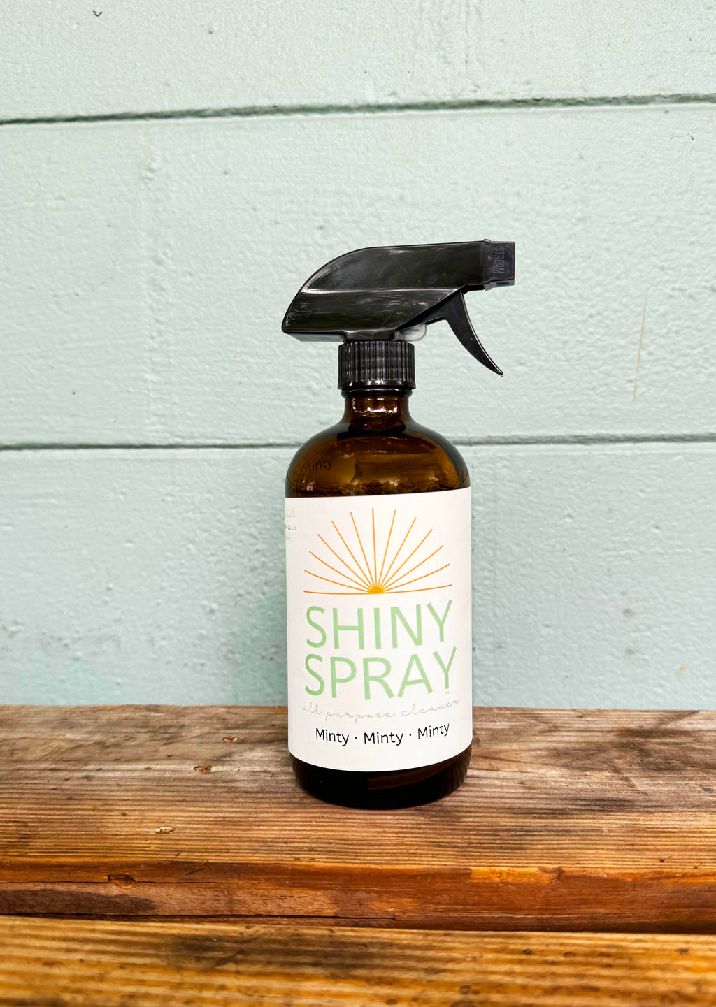 Brown spray bottle labeled 'Shiny Spray' on a wooden surface with a light blue wooden panel background.