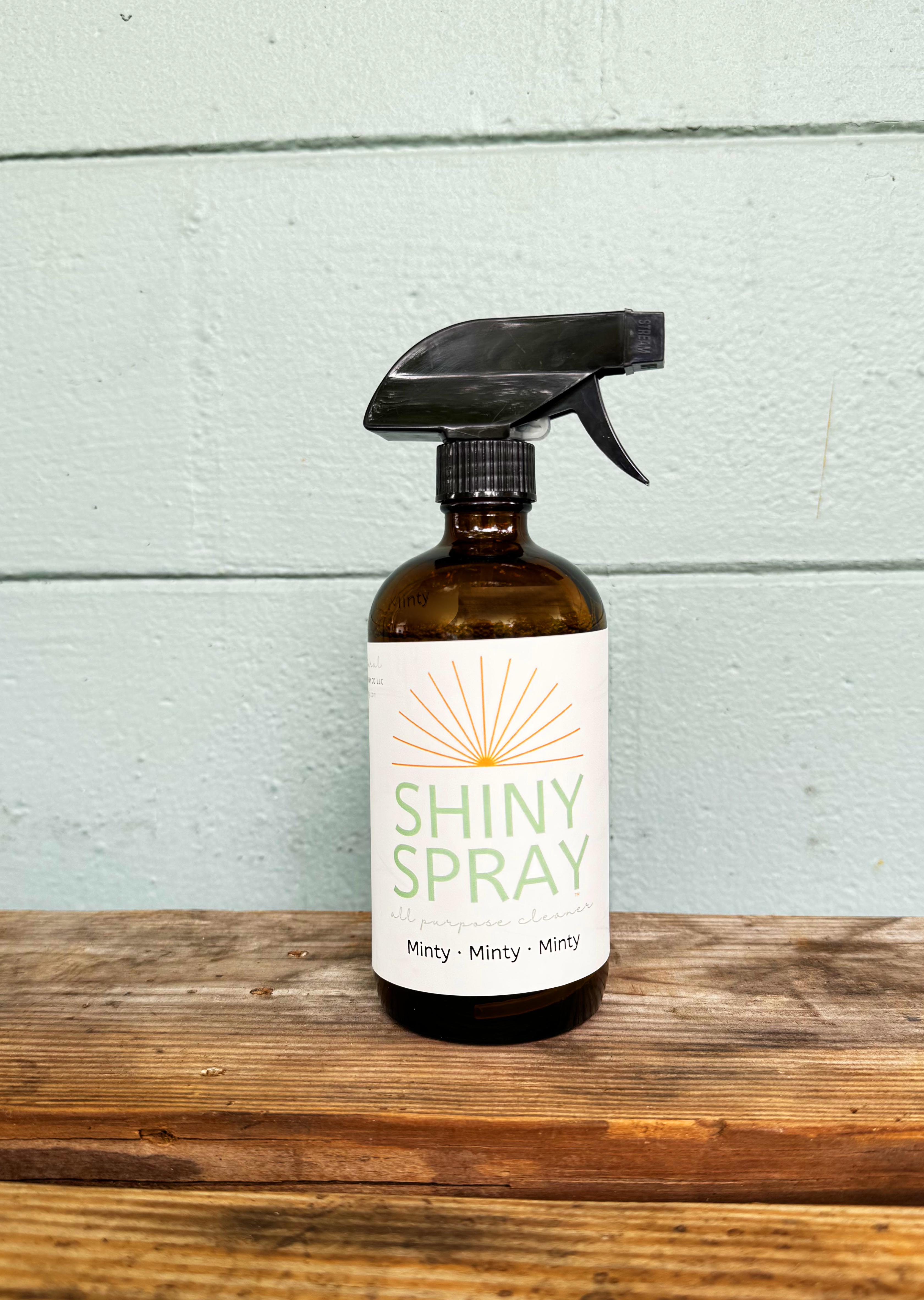 Brown spray bottle labeled 'Shiny Spray' on a wooden surface with a light blue wooden panel background.